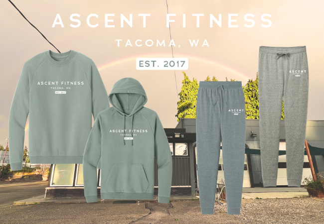 Four pieces of Ascent Fitness Tacoma, WA apparel—crewneck, hoodie, joggers, and sweatpants—are displayed in front of a gym building with a rainbow and sunset sky in the background. Text says EST. 2017..